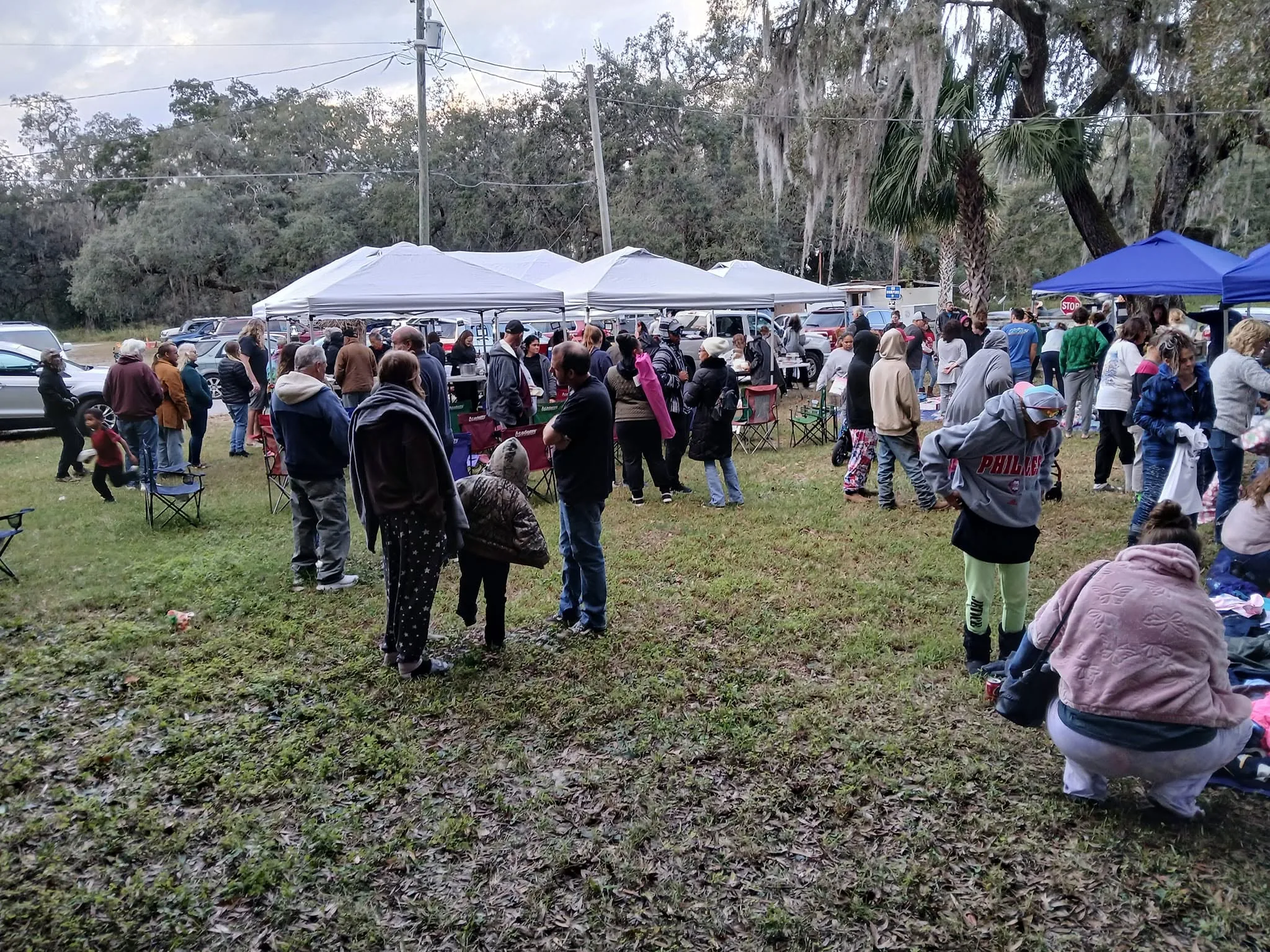 A large crowd gathered outdoors under oak trees at a WOTS outreach
